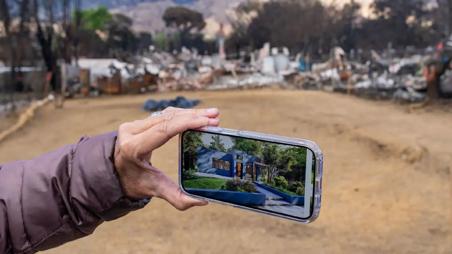 A hand holds a phone showing a house rendering in front of a cleared lot and damaged structures.
